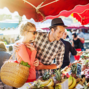 a young couple buying fruits and vegetables in a market on a sunny morning, the young woman carries a basket